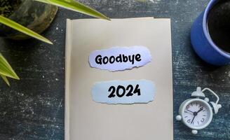 Goodbye 2024 text in notebook with alarm clock on wooden table. photo