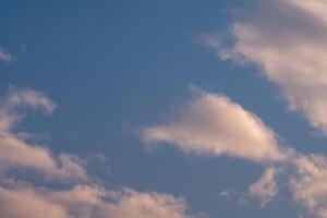 Soft fluffy clouds drift across a serene blue sky bathed in the warm glow of the setting sun photo