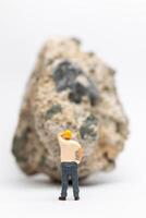 Miniature people construction worker stands next to a massive rock scratching his head in contemplation photo