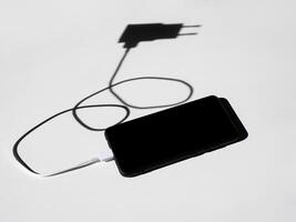 black smartphone with a charging wire on a white table with shadows. photo