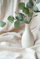 Styled display features a matte white ceramic bottle filled with fresh eucalyptus leaves, elegantly placed on soft beige linen fabric, providing ample space for text overlay and branding photo