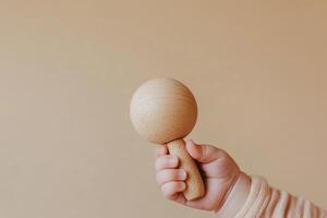 A baby's delicate hand holds a wooden rattle, showcasing a minimalist design against a smooth beige backdrop. Soft shadows enhance the simplicity and elegance of the moment photo