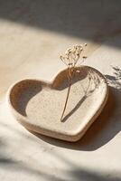 This heart-shaped plate made of light stone rests on a smooth neutral surface. A small dried flower adds a gentle accent, creating a serene and inviting atmosphere perfect for text overlay photo