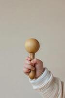 A baby holds a smooth wooden rattle with a firm grip, set against a calming beige background. The gentle shadows and soft tones create a serene and minimalist atmosphere photo