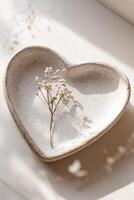 Heart-shaped plate crafted from light stone rests on a neutral surface, showcasing a delicate dried flower accent. The soothing ambiance invites creativity for text overlay photo