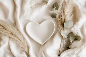Heart-shaped plate in matte white ceramic sits on soft beige linen, surrounded by dried pampas grass and eucalyptus leaves, creating a cozy and inviting natural arrangement photo