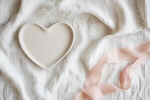 A minimalist heart-shaped plate in light taupe rests on a soft linen tablecloth, accompanied by a delicate blush pink ribbon, creating a cozy and refined atmosphere suitable for any occasion photo