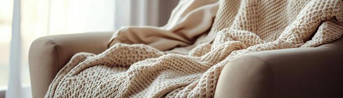 A soft beige knitted blanket is casually draped over a stylish armchair in a well-lit living room. Sunlight filters through sheer curtains, creating a warm and inviting atmosphere photo