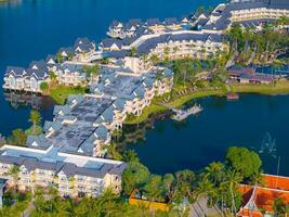 Aerial view of a resort with gray roofed buildings, lush greenery, a lagoon, a dock, and a fountain, set in a tropical coastal setting in Phuket, Thailand. photo