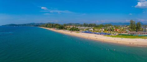 Aerial view of a long sandy beach in Phuket, Thailand, with turquoise waters, lush greenery, resort buildings, and rows of blue umbrellas along the shore. photo