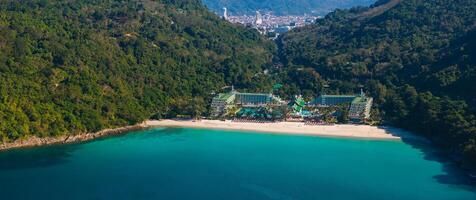 Aerial view of a secluded beach with turquoise waters, lush green hills, and a large resort complex. A distant urban area is visible in the background. photo