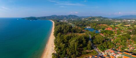 Aerial view of a sandy beach in Phuket, Thailand, bordered by turquoise waters, a lotus pond, red roofed buildings, and rolling hills in the distance. photo
