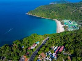 Aerial view of a crescent shaped beach in Phuket, Thailand, with turquoise waters, lush forest, colorful buildings, and a resort near the shoreline. photo