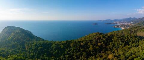 Aerial view of a lush green forest leading to a calm blue sea, with a mix of natural greenery, buildings, a small bay, and a clear horizon in Thailand. photo