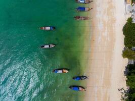 Aerial view of a serene beach with turquoise waters, colorful long tail boats anchored along the shore, and lush green trees framing the scene. photo
