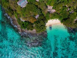 Aerial view of a small beach surrounded by lush vegetation and rocky coastline in Thailand. Turquoise waters reveal coral, with boats near the shore. photo