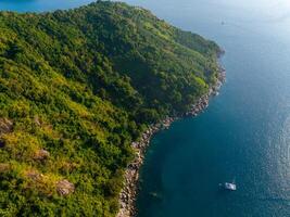 Aerial view of a lush, green coastline with rocky shores, clear blue waters, and a single white yacht near Phuket, Thailand's hilly terrain. photo