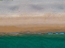 Aerial image of a beach with turquoise ocean waters, wet sandy shoreline, and dry textured sand. Sparse greenery is visible at the edge of the frame. photo