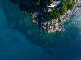 Aerial image of a rocky coastline with turquoise waters, lush greenery, and a white structure with a red tiled roof in Phuket or Phi Phi Islands. photo