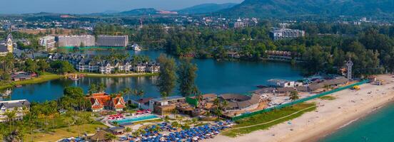 A sandy beach in Phuket, Thailand, with colorful umbrellas, a calm lagoon, lush greenery, and a red roofed Thai style building near urban hills. photo