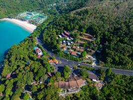 Aerial perspective of Phuket, Thailand, showing a turquoise beach, dense greenery, a winding road, and buildings nestled among tropical vegetation. photo