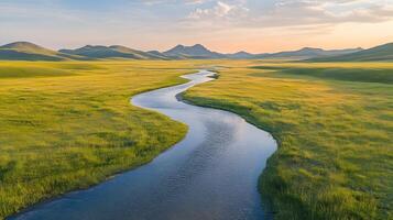 Pristine meandering river flows through untouched green meadows with rolling hills and majestic mountain backdrop, captured in aerial view during hour, showcasing vibrant spring colors. photo