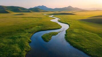 Pristine meandering river flows through untouched green meadows with rolling hills and majestic mountain backdrop, captured in aerial view during golden, showcasing vibrant spring colors. photo