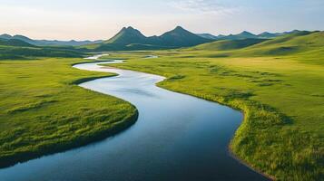 Pristine meandering river flows through untouched green meadows with rolling hills and majestic mountain backdrop, captured in aerial view during golden hour, vibrant spring colors. photo