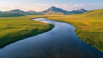 Pristine meandering river flows through untouched green meadows with rolling hills and majestic mountain backdrop, captured in aerial view during golden hour, showcasing vibrant spring photo