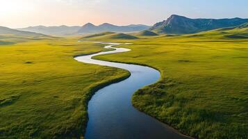 Pristine meandering river flows through untouched green meadows with rolling hills and majestic mountain backdrop, captured in aerial view during golden hour, showcasing vibrant colors. photo