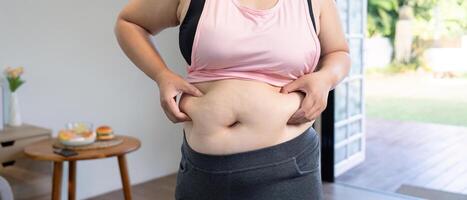 Woman gripping her waist, reflecting on body image and lifestyle choices while standing in a bright room photo