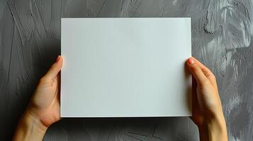 A person holds a blank white paper sheet in front of a grey concrete wall photo