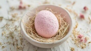 Pink decorative egg resting in a bowl surrounded by dried flowers and straw on a light surface photo