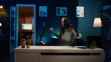 Girl in neon lit apartment using phone on selfie stick to capture welcoming intro for vlogging channel. Joyful entertainer in living room connecting with loyal audience during live stream, camera B video