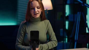 Young content creator records introductory with smartphone, waving hand, inviting viewers to subscribe to channel. Media star uses mobile phone to film herself connecting with audience, camera A video