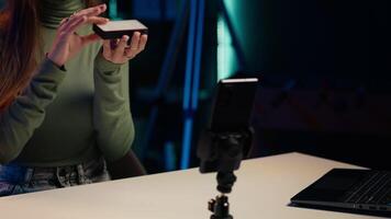 Gen Z girl in apartment studio using mobile phone to record, reviewing mini RGB lights. Young entertainer films electronics haul with cellphone on tripod, presenting LED lighting device, camera A video