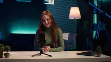 Smiling girl using smartphone on tripod to film herself discussing topics of interest for generation Z fanbase. Influencer recording targeting gen Z viewers using phone selfie camera, camera A video