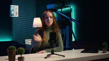 Smiling girl using smartphone on tripod to film herself discussing topics of interest for generation Z fanbase. Influencer recording targeting gen Z viewers using phone selfie camera, camera B video