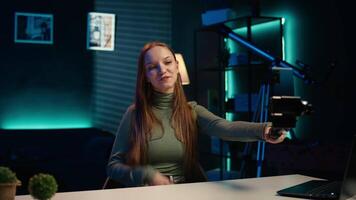Upbeat teenager films content for online streaming channel in apartment studio using phone on selfie stick. Smiling gen z influencer at home interacts with subscribers, recording with phone, camera B video