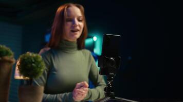 Young content creator records introductory with smartphone, waving hand, inviting viewers to subscribe to channel. Media star uses mobile phone to film herself connecting with audience, camera B video
