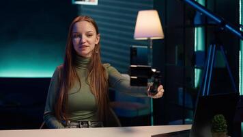 Girl in neon lit apartment using phone on selfie stick to capture welcoming intro for vlogging channel. Joyful entertainer in living room connecting with loyal audience during live stream, camera A video