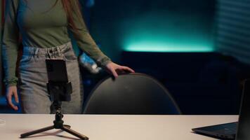 Happy girl arriving in studio, filming with smartphone on tripod, discussing topics of interest for gen Z fanbase. Jolly media star entering home studio, recording with mobile phone, camera A video