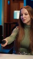 Vertical Upbeat teenager films content for online streaming channel in apartment studio using phone on selfie stick. Smiling gen z influencer at home interacts with subscribers, recording with phone, camera A video