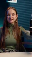 Vertical Girl in neon lit apartment using phone on selfie stick to capture welcoming intro for vlogging channel. Joyful entertainer in living room connecting with loyal audience during live stream, camera A video