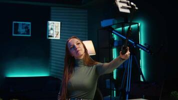 Gen Z teenager recording introductory with cellphone on selfie stick, inviting viewers to subscribe to channel. Influencer in home studio uses selfie camera to film herself, camera B video