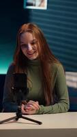 Vertical Smiling girl using smartphone on tripod to film herself discussing topics of interest for generation Z fanbase. Influencer recording targeting gen Z viewers using phone selfie camera, camera A video