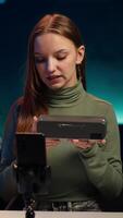 Vertical Influencer assessing portable speaker performance, talking about parts used, filming with phone. Girl recording music device review using smartphone on tripod, pleased with used materials, camera A video