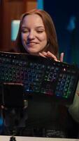 Vertical Gen Z girl presenting RGB lit mouse and keyboard while doing review in personal studio to see if price is justified. Young influencer showcasing neon lit computer peripheral features to fans, camera A video