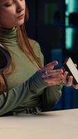 Vertical Cheerful gen Z girl in home studio reviewing mini RGB lights controller, using microphone to record. Smiling influencer films electronics haul, presenting LED lighting device, camera A video