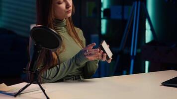 Cheerful gen Z girl in home studio reviewing mini RGB lights controller, using microphone to record. Smiling influencer films electronics haul, presenting LED lighting device, camera A video